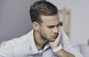 A person wearing a light blue shirt and white watch is holding their cheek with a pained expression, possibly experiencing toothache.