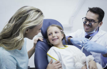 A child sits in a dental chair, smiling at a woman beside her, while a dentist with a dental mirror smiles nearby, wearing gloves and a mask.