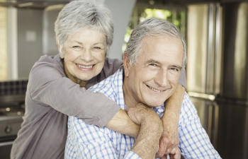 Older couple smiling, with the woman hugging the man from behind, inside a kitchen.