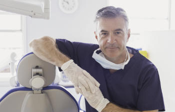 A dentist in scrubs and gloves stands beside a dental chair in a bright office, looking at the camera.