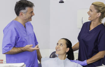 A dental professional and an assistant engage in conversation with a seated patient in a dental office.