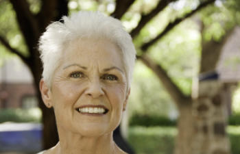 Elderly woman smiling outdoors with blurred trees in the background.