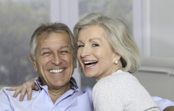 An older couple is sitting and smiling cheerfully indoors, with a blurred background.
