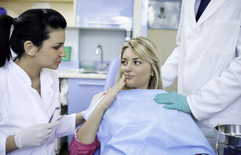 A patient sits in a dental chair with a dental assistant and a dentist standing beside her in a clinic setting.