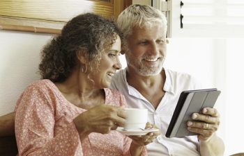 An older couple sits on a couch beside a window, smiling. The woman holds a cup while the man holds a tablet.