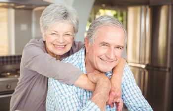 An older couple smiling in a kitchen. The woman is embracing the man from behind.