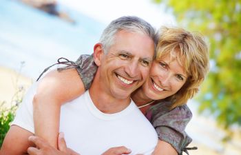 A couple smiling, the woman embracing the man from behind. They are outdoors near the beach, with blurred greenery and ocean in the background.