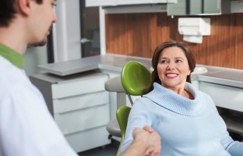 A woman in a dental chair smiles and shakes hands with a dentist in a modern dental office.