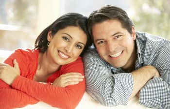 A smiling couple leans on a white surface, both resting their arms in front. The woman wears a red top, and the man wears a checkered shirt.