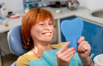 Woman with red hair smiling and sitting in a dental chair, holding a tooth-shaped mirror in a dental clinic setting.