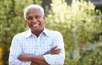 Smiling man with gray hair and a plaid shirt stands outdoors with arms crossed, blurred greenery in the background.