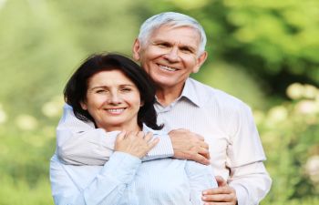 An older couple stands outside, smiling. The man embraces the woman from behind. The background is a blurred green landscape.