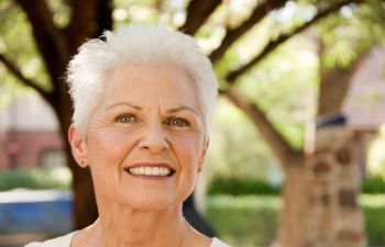 An older woman with short white hair smiles outdoors, standing in front of trees and blurred foliage.