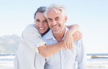 A smiling couple embraces on a sunny beach with waves and distant hills in the background.