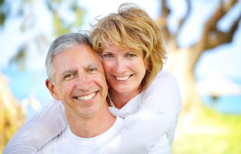Smiling couple outdoors, woman embracing man from behind against a blurred green and blue background.