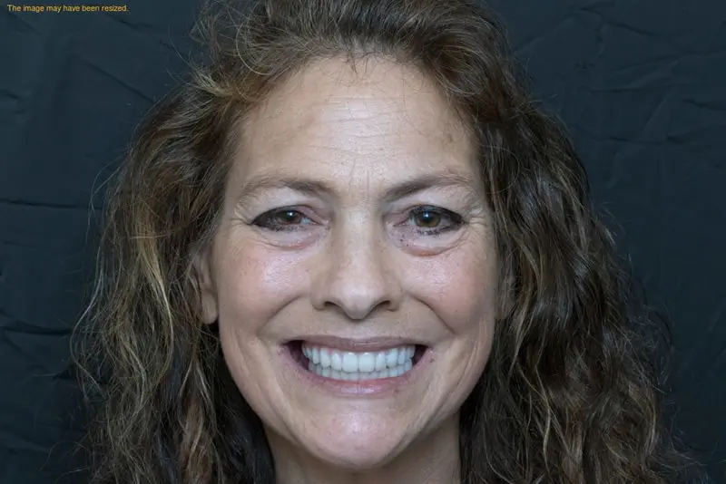 Woman with curly hair, smiling broadly, stands in front of a dark background.