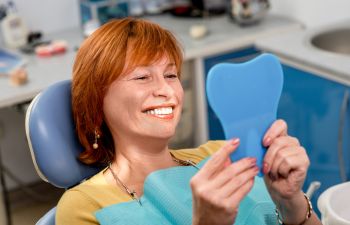 Woman sitting in a dental chair, smiling and holding a tooth-shaped mirror.