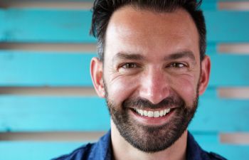 Smiling man with a beard and dark hair, in front of a turquoise wooden wall.
