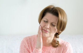A woman with light brown hair is touching her cheek with a pained expression, possibly indicating a toothache. She is wearing a pink sweater.