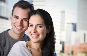 A man and woman smiling, standing closely together with a blurred cityscape in the background.