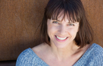 A woman with shoulder-length brown hair and bangs is smiling at the camera. She is wearing a blue knitted sweater. The background is a textured brown surface.