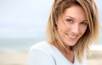 Woman with short blonde hair smiling slightly, wearing a light-colored sweater, standing outdoors near a beach with the ocean in the background.