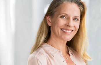 Smiling woman with long blonde hair, wearing a light blouse, against a soft, light background.