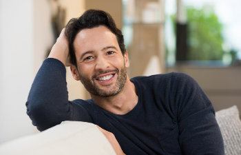 A man with dark hair and a beard, wearing a black sweater, leans on a light-colored couch and smiles at the camera.