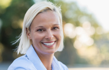 Smiling person with short blonde hair outdoors, wearing a light blue shirt, with trees blurred in the background.
