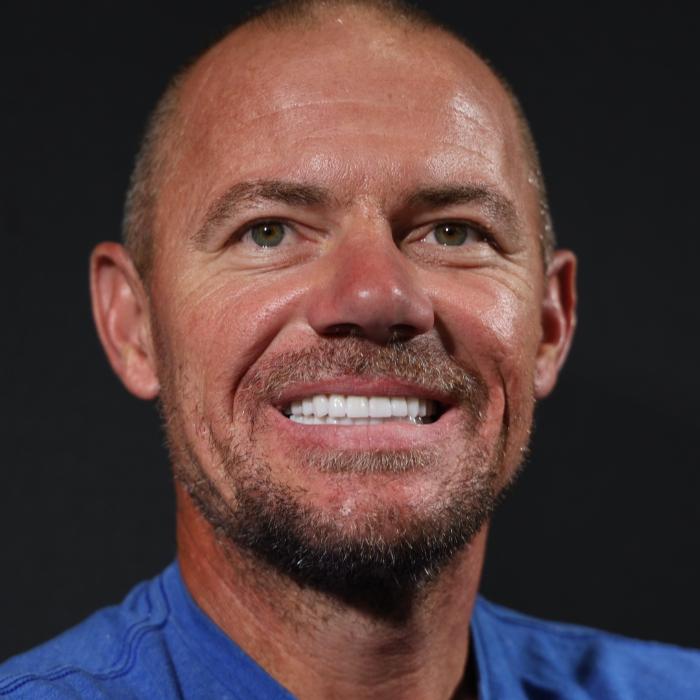 A man with short hair and a beard smiles at the camera against a dark background. He is wearing a blue shirt.