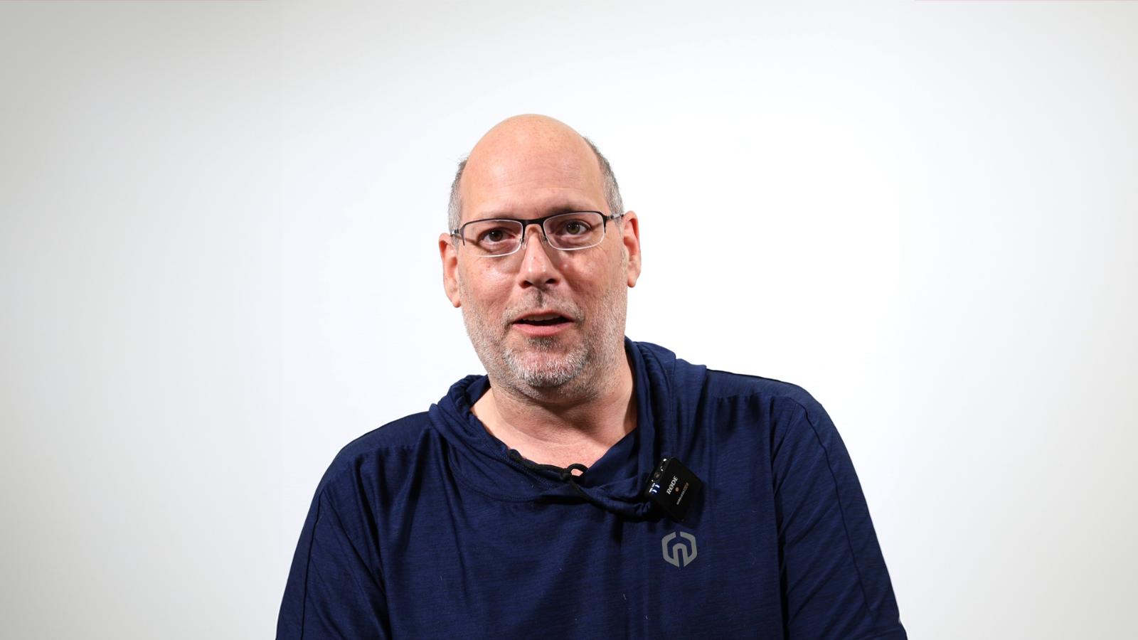 A bald man wearing glasses and a dark blue hoodie is speaking against a plain white background.