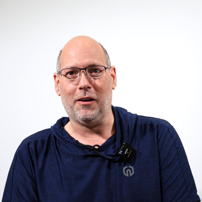 A man with glasses and a blue hoodie stands against a plain white background, looking at the camera.