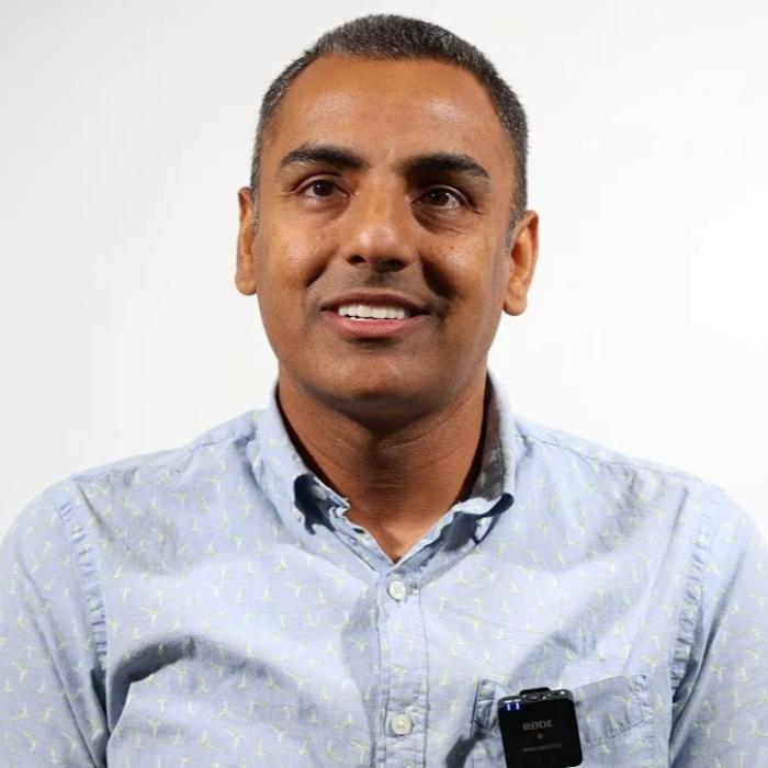 Person wearing a light blue shirt with a microphone attached, smiling, against a plain background.