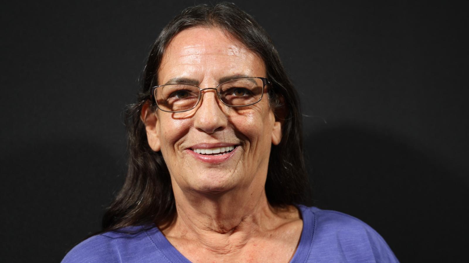 Woman with long brown hair and glasses smiles against a dark background, wearing a purple top.