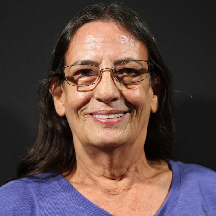A woman with long dark hair and glasses smiles at the camera in front of a black background, wearing a purple top.