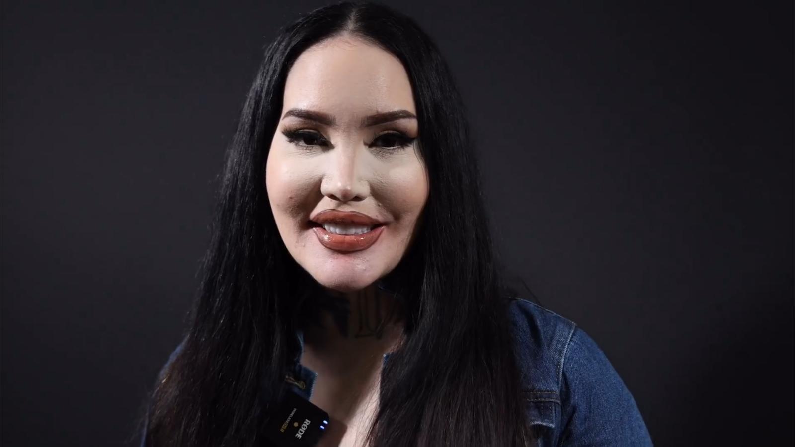 A person with long black hair and a denim jacket stands against a dark background, smiling softly at the camera.