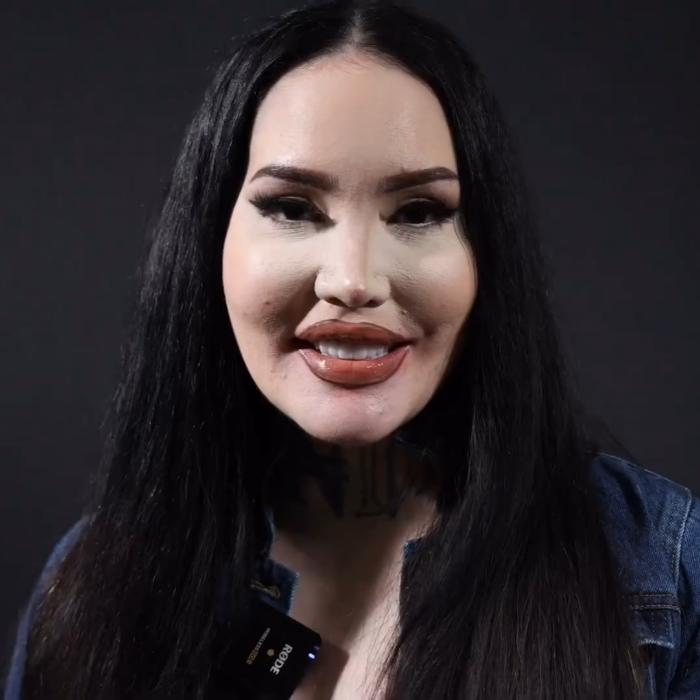 Person with long black hair, wearing a dark top, smiling in front of a plain dark background.