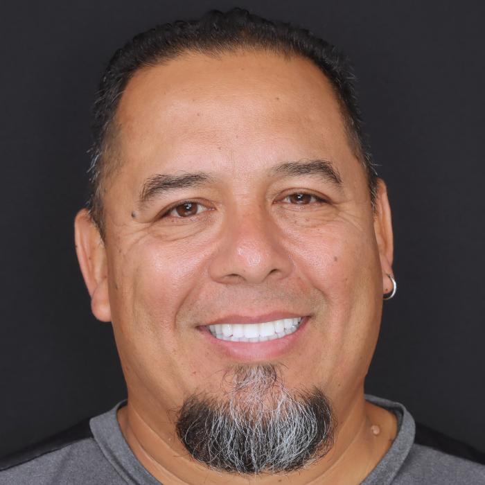 Smiling man with a short beard and an earring, wearing a gray and black shirt, against a black background.