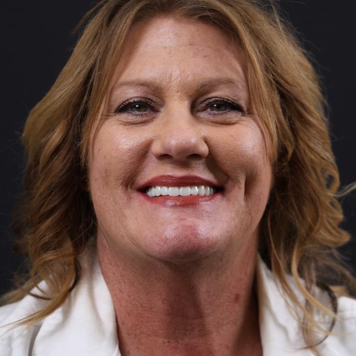 Smiling person with wavy hair wearing a light-colored top against a dark background.