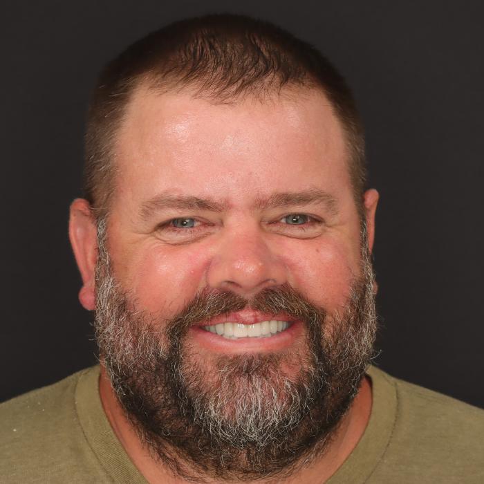Man with a beard and short hair, smiling against a black background, wearing a green shirt.