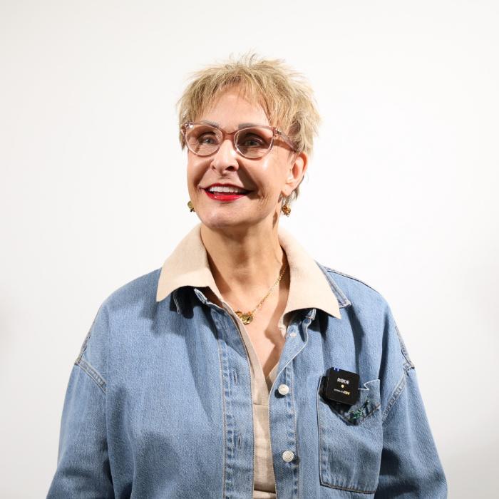 Person with short hair and glasses wearing a denim jacket over a beige shirt. They are looking to the side and smiling against a plain background.