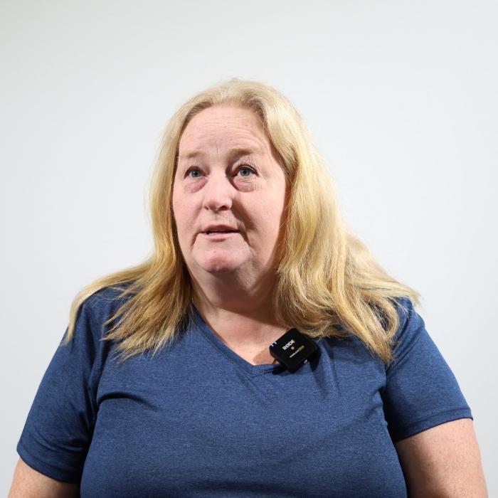 A woman with long blonde hair wearing a blue shirt and a small microphone clipped to her collar sits against a plain white background.
