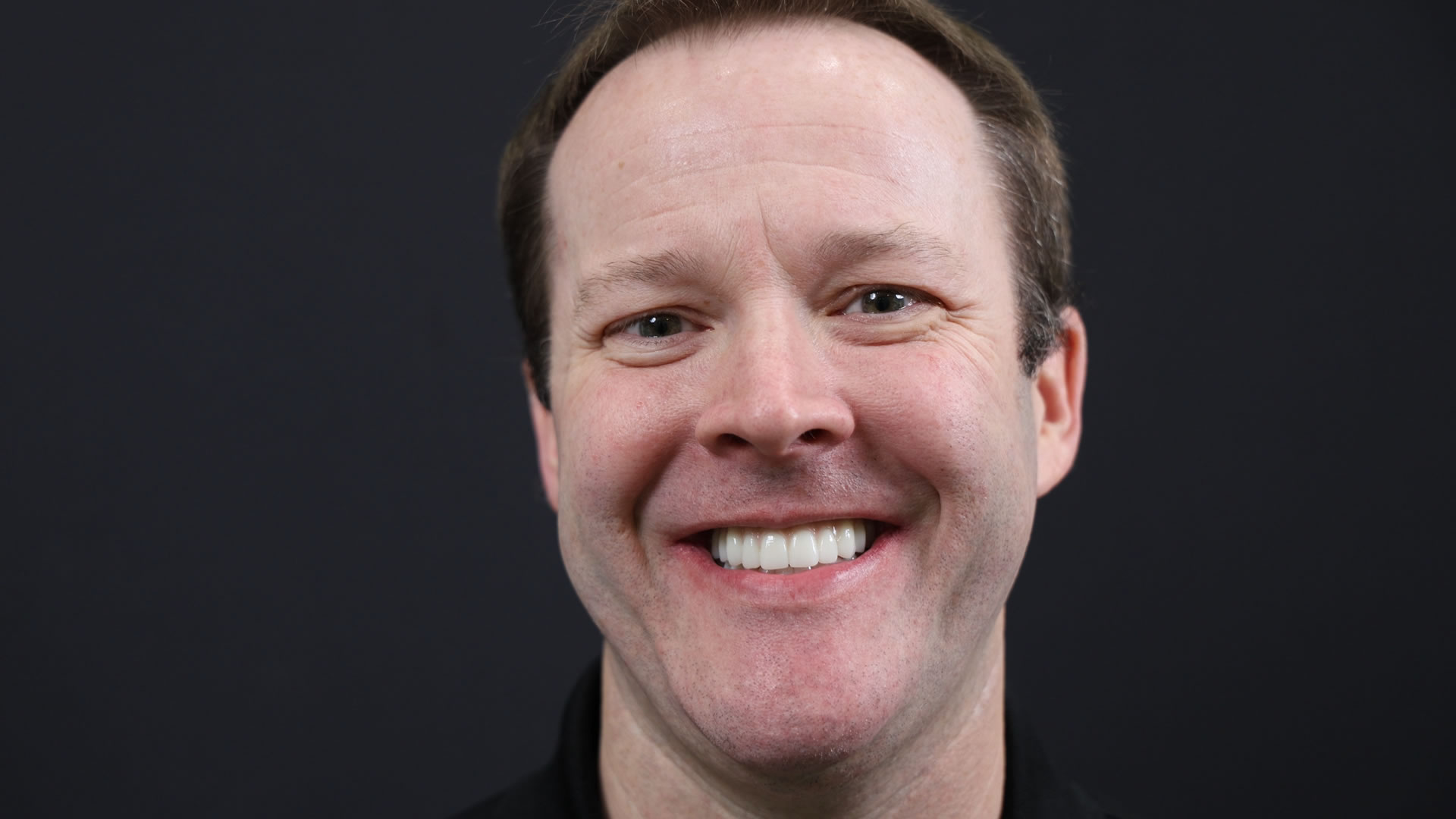 Smiling man with short hair and a black shirt against a dark background.
