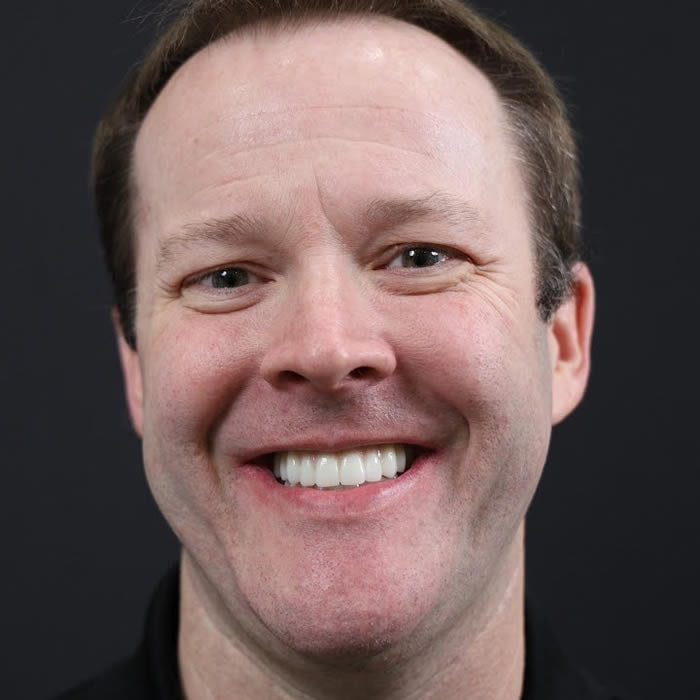 A man with short hair smiles broadly against a dark background.