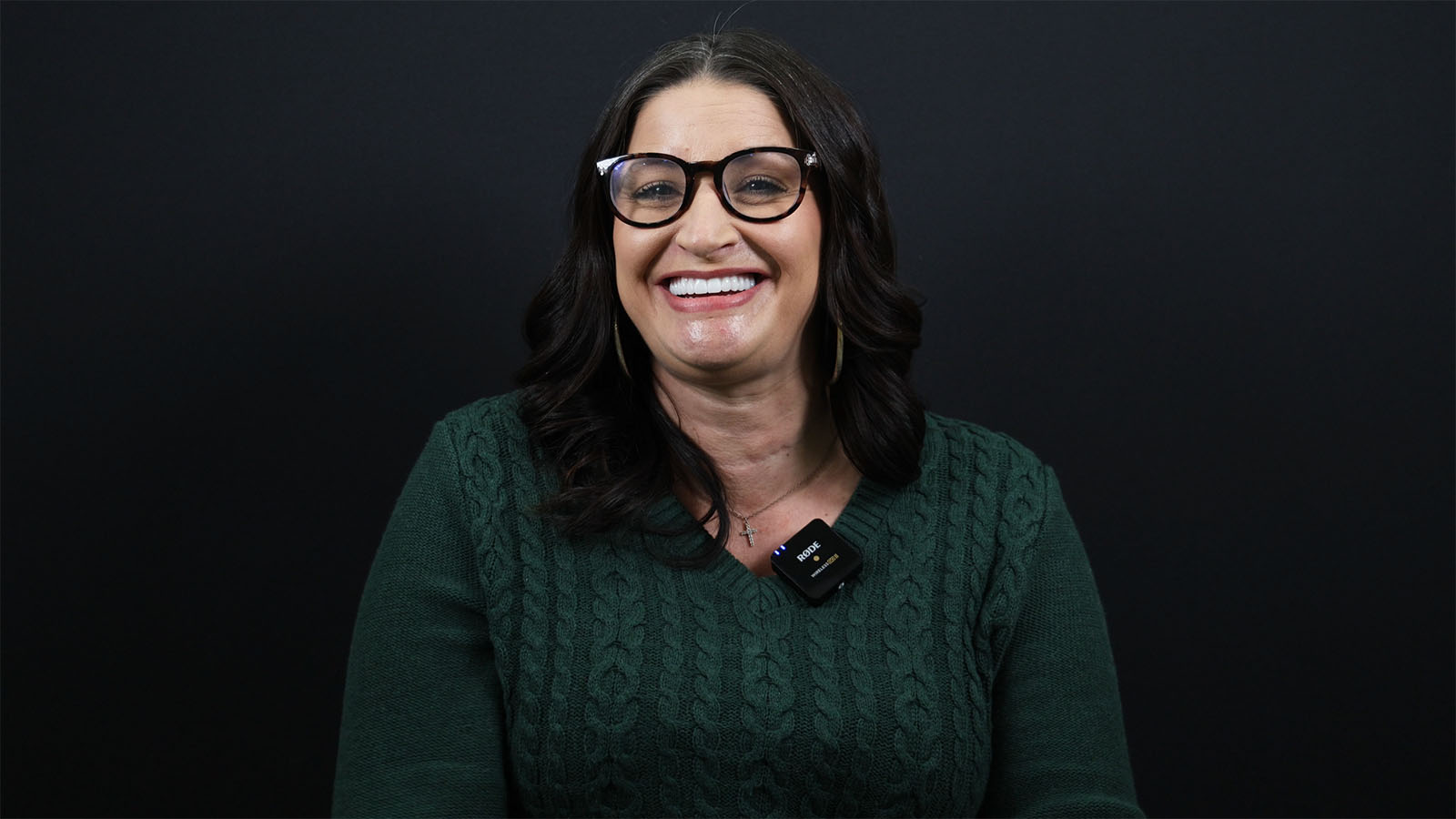 A woman with long dark hair and glasses smiles, wearing a green sweater and a microphone against a black background.