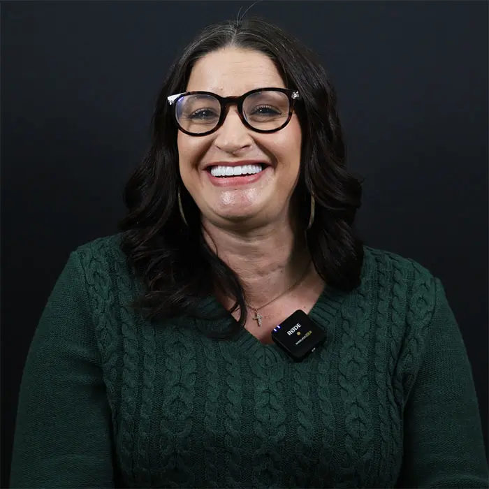 Woman with glasses wearing a green sweater and a microphone smiles against a black background.
