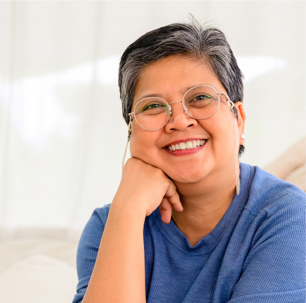 Person with short hair and glasses, wearing a blue shirt, smiling with hand under chin against a soft-focus light background.
