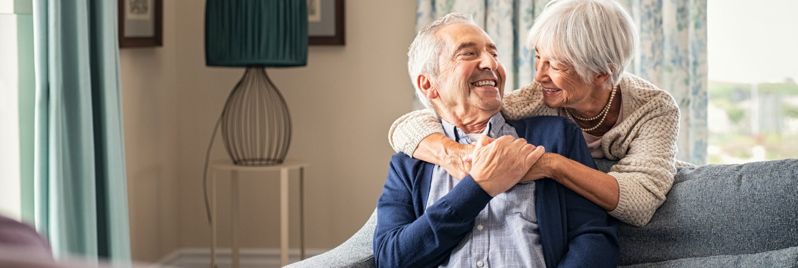 Elderly couple sitting on a couch smiling and embracing warmly in a cozy living room setting.