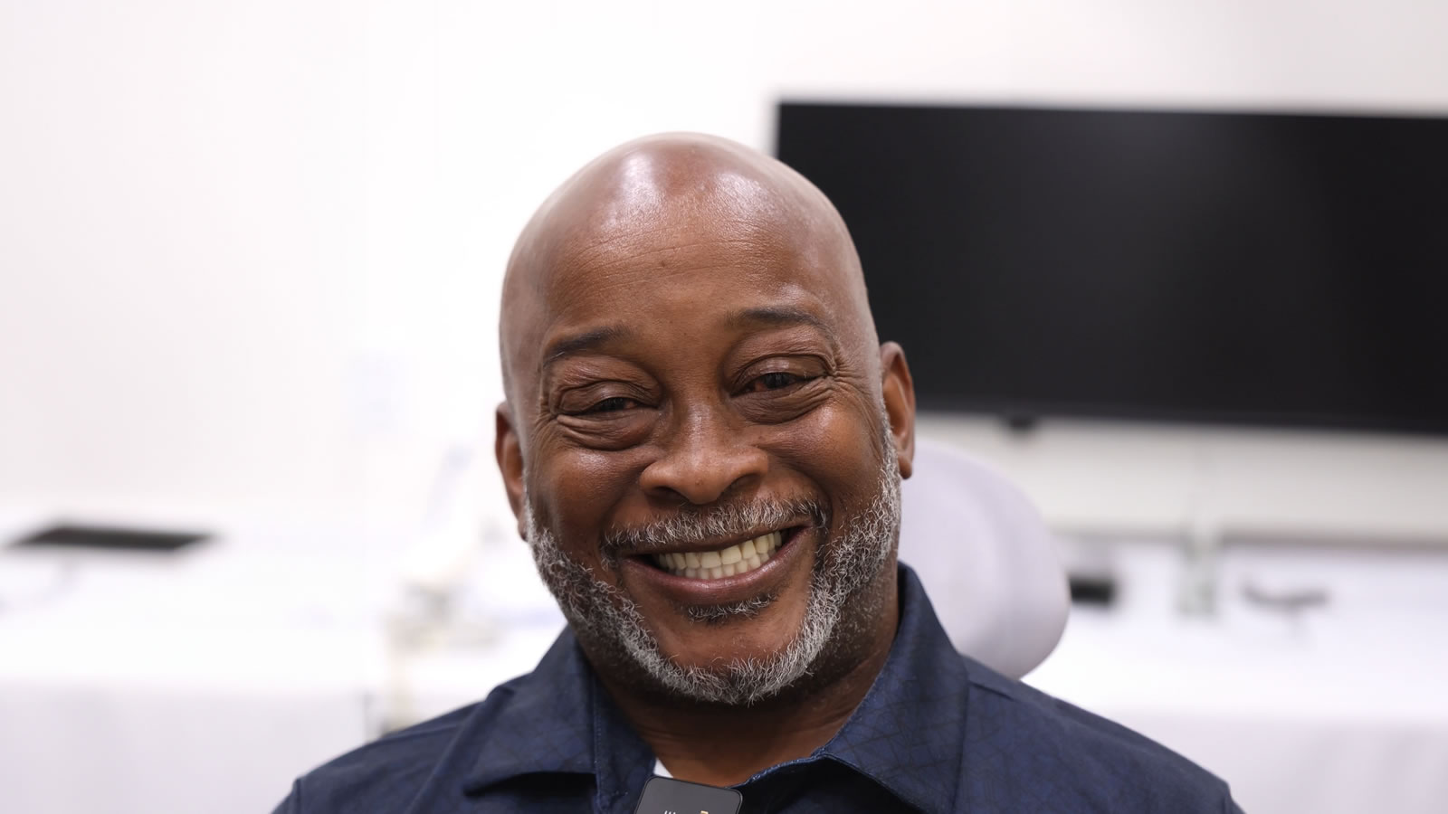 A man with a shaved head and gray beard smiles while seated indoors, with a blurred monitor and white background behind him.