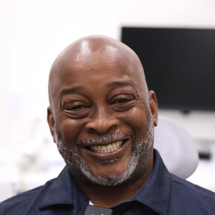 A middle-aged man with a bald head and gray beard smiles while sitting indoors, with a monitor and office equipment in the background.
